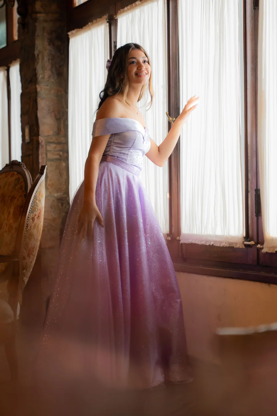 Quinceañera con vestido lila junto a la ventana